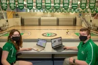 Two sport media students watching over the basketball court with their laptops in front of them, looking back at the camera, smiling for a photo 