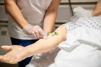 A close-up image shows a nursing student's gloved hands inserting an IV into the arm of a practice mannequin in the nursing simulation lab. 