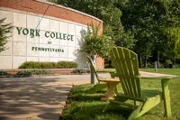 A large sign shows the York College of Pennsylvania logo on a brick wall as two green Adirondack chairs sit in the foreground on an autumn day in the campus quad 