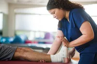 A physical therapist wearing navy blue scrubs stretches a patient's foot that is wrapped in an ace bandage. 
