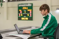 A student wearing a green and white track jacket types at a laptop in a gymnasium. A scoreboard is visible in the background. 