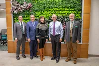 A group of five including visiting state government representatives, President Thomas Burns, and faculty pose in front of a wall of plants in the Appell Horticulture Center. 