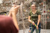 A student smiles as she prepares herself with ropes and harness to climb the rock wall in the Grumbacher Center. Her spotter is visible, blurred in the foreground.