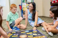 A teacher sits on the floor with three of her students, pointing at images of the solar system on a play mat.