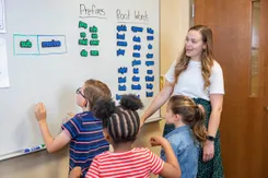 A teacher stands by a whiteboard in a classroom and watches as three students work with magnets featuring prefixes and root words.