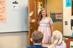 A teacher enters an elementary school classroom, smiling and waving at her seated students who are visible in the foreground.
