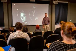 A professor speaks to a group of students sitting in the film viewing room with black and white film on the screen over his shoulder.