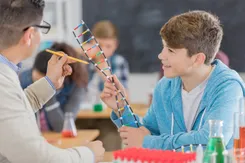 A high school student holds a plastic model of a DNA helix as his teacher looks on, pointing at areas of the model with his pencil.