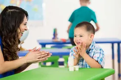 A teacher and an elementary student clap in front of a pile of blocks in a colorful classroom full of plastic kid-sized tables.