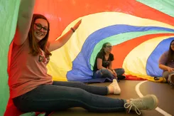 College students sit under the rainbow colors of a recreational parachute, smiling as the fabric billows around them.