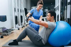 A trainer wearing scrubs assists a young man pulling pulley weights while he leans against a therapy ball in a gym.