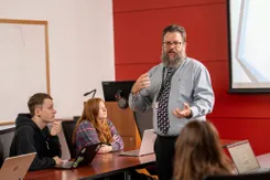 A professor in a button-down shirt and tie lectures in front of a group of students in a classroom with a bright red wall.