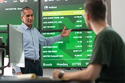 Professor Pawan Madhogarhia points to a large touch-screen in the Willman Business Center's NASDAQ lab. A student looks on.