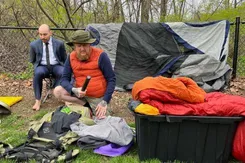 A photo from a scene during an outdoor live play shows two men sitting amid camping gear. One is dressed for hiking; the other wears a suit and is barefoot.