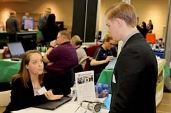 A student stops at a job fair table to speak with a potential employer. Both wear business suits.