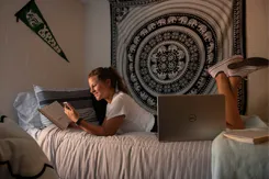 A student lounges on her dorm room bed, reading a book and glancing at her cell phone.