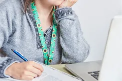 A young woman rests her chin in her hand while taking notes and looking at a laptop. She wears a sunflower lanyard.