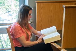 A student sits at a study booth in the library, reading from a large hardcover book.