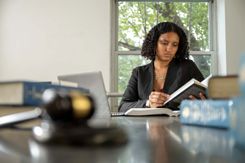 A student sits at a table in the library, reading with a stack of books and her laptop in front of her. She wears a suit jacket and trees are visible through the window behind her.