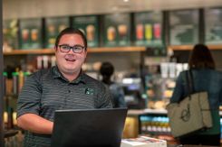 A student smiles at someone off-camera while working on his laptop in the campus Starbucks.