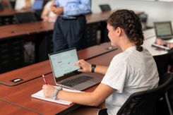 A student sits at a group desk, pointing at her laptop and writing in a notebook.