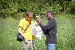 Two students look at a sample container while in a green field, surrounded by trees in the backdrop