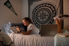 A student lounges on her dorm room bed, reading a book and glancing at her cell phone.