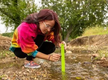 A student crouches by the edge of the creek, measuring water levels.