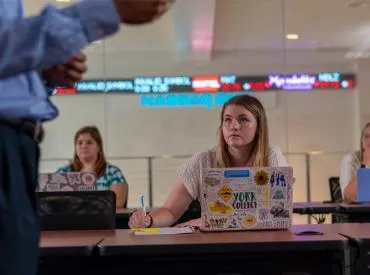 A student looks on as a professor lectures in the foreground. A NASDAQ ticker scrolls across the wall behind her desk.