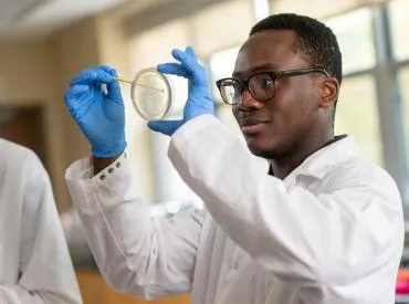 A student swabs a petri dish in a lab, wearing glasses and protective gloves and white lab coat
