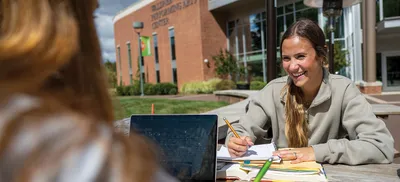 Two students sit cheerfully at a picnic table outside the Performing Arts Center, study materials spread on the table in front of them.