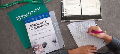 A photo of a tabletop shows a students hands writing on a piece of torn notebook paper alongside a tablet, a York College branded folder and lanyard, and an Introduction to Entrepreneurship textbook