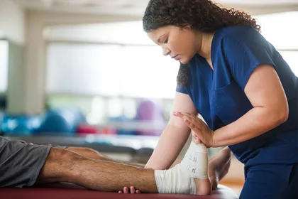 A physical therapist wearing navy blue scrubs stretches a patient's foot that is wrapped in an ace bandage. 