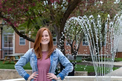A YCP student posing in front of the campus fountain in a pink dress and blue jean jacket 