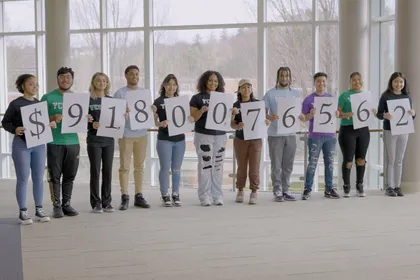 A group of students stand in line, holding printed numbers that spell out $91,800,765.62 