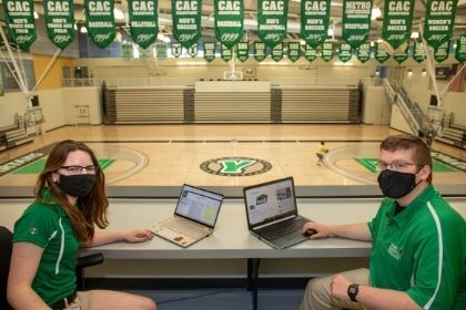 Two sport media students watching over the basketball court with their laptops in front of them, looking back at the camera, smiling for a photo