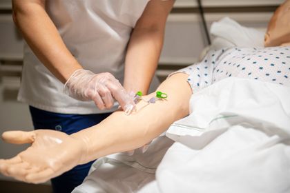 A close-up image shows a nursing student's gloved hands inserting an IV into the arm of a practice mannequin in the nursing simulation lab.