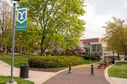 The York College campus quad is shown in part, with a brick pathway leading up to the Performing Arts Center on a spring day. Picnic tables line the path and a York College banner stands proudly from its place on a light post.