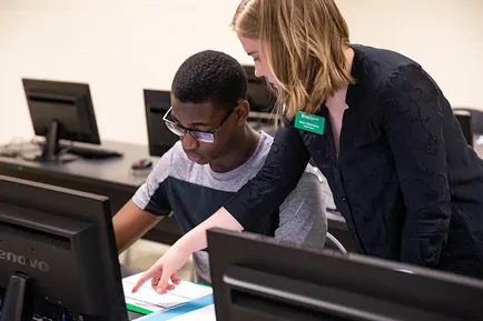 A staff member stands beside a student seated in the computer lab, pointing at paperwork in front of him as he references his screen.