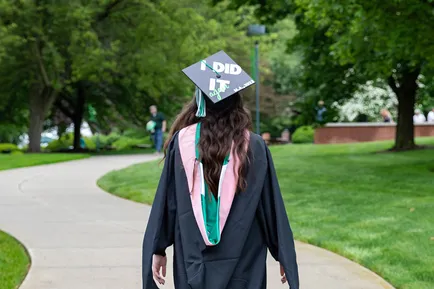 A graduating student walks across campus in her regalia, facing away from the camera. The top of her cap is visible and decorated to read 