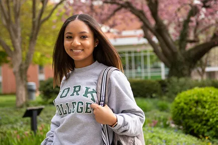 A student wearing a York College sweatshirt smiles at the camera, backpack slung over one shoulder. Springtime flowers and trees bloom behind her.