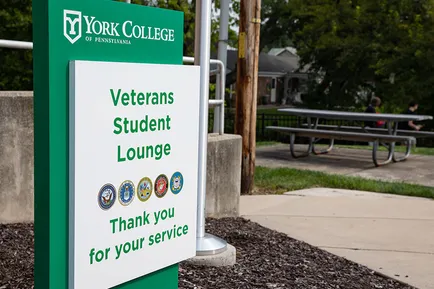 A sign with the York College logo at the top sits outside a campus building. It reads: Veterans Student Lounge. Thank you for your service.