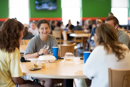 Four students gather around a dining hall table, chatting with their meals in front of them. 