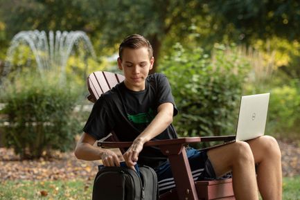 A student pulls his laptop from a backpack as he sits in an Adirondack chair in front of the fountain on the campus quad