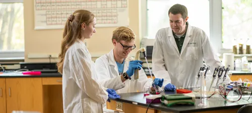Two students and a professor wear lab coats and safety gloves while using equipment in a biology laboratory.