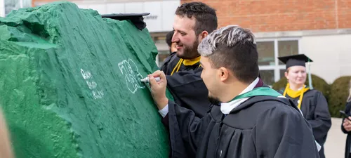 A graduate wearing regalia signs the green rock at the center of campus.