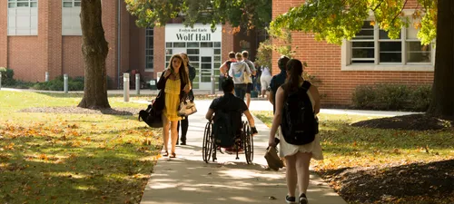 A group of students, one using a wheelchair, head to and from class in Wolf Hall on an autumn day with leaves falling on the campus quad around them.