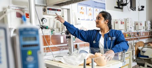 A student nurse reaches for a monitor in the simulation lab, surrounded by medical equipment and a 