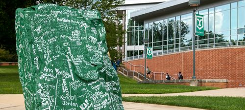 The rock, painted green and signed with graduated student signatures, sits in front of campus buildings