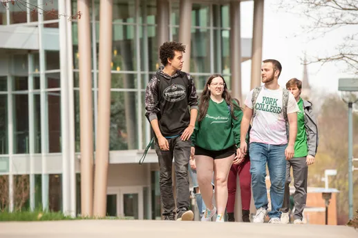 A group of students talk and laugh while walking up the sidewalk in front of the Performing Arts Center grand glass entryway.